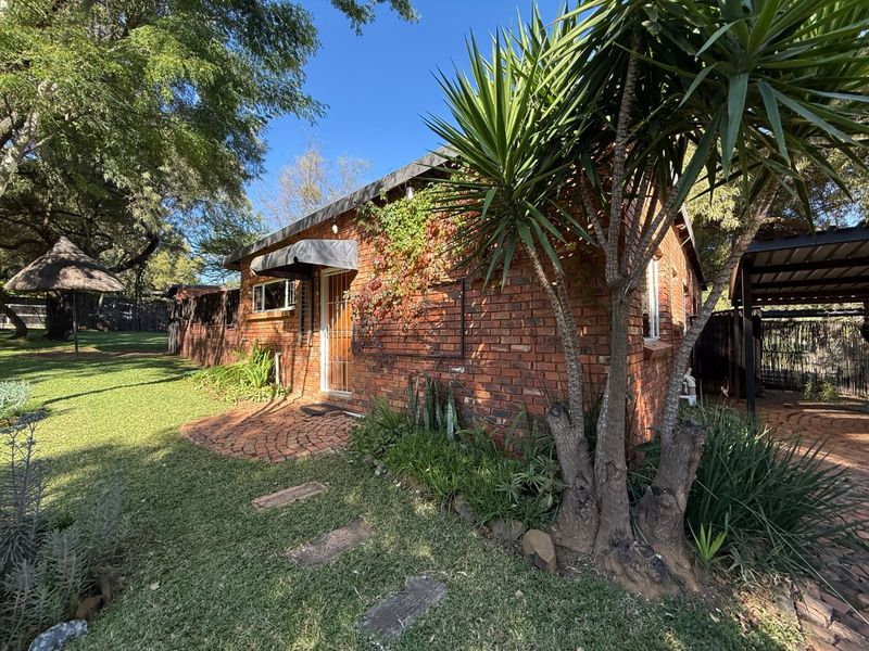 Rear view of the face brick house featuring a paved patio area, security entrance, and adjacent carport set within a landscaped garden.