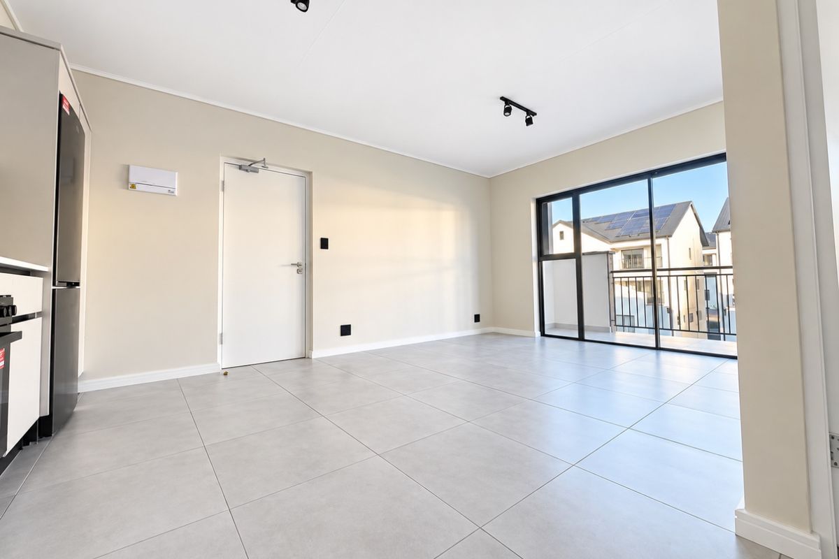 A bright, modern open-plan living area featuring large format floor tiles, track lighting, and a sliding glass door leading to a private balcony.