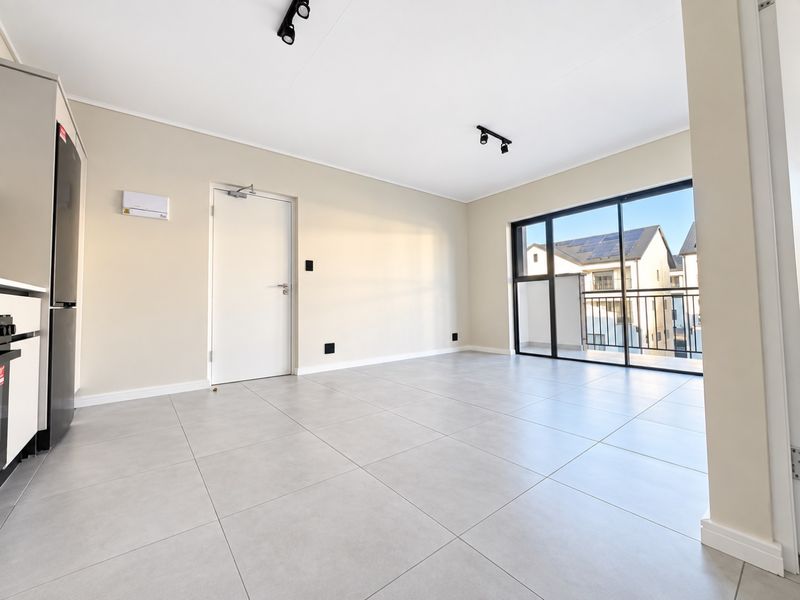 A bright, modern open-plan living area featuring large format floor tiles, track lighting, and a sliding glass door leading to a private balcony.
