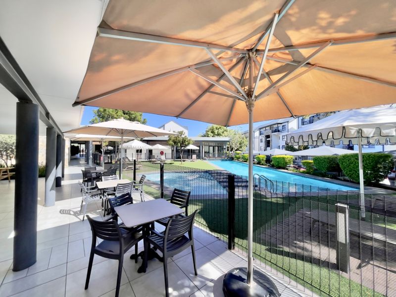 A well-maintained communal pool area featuring a tiled patio, safety fencing, and landscaped garden surroundings, ideal for outdoor relaxation.