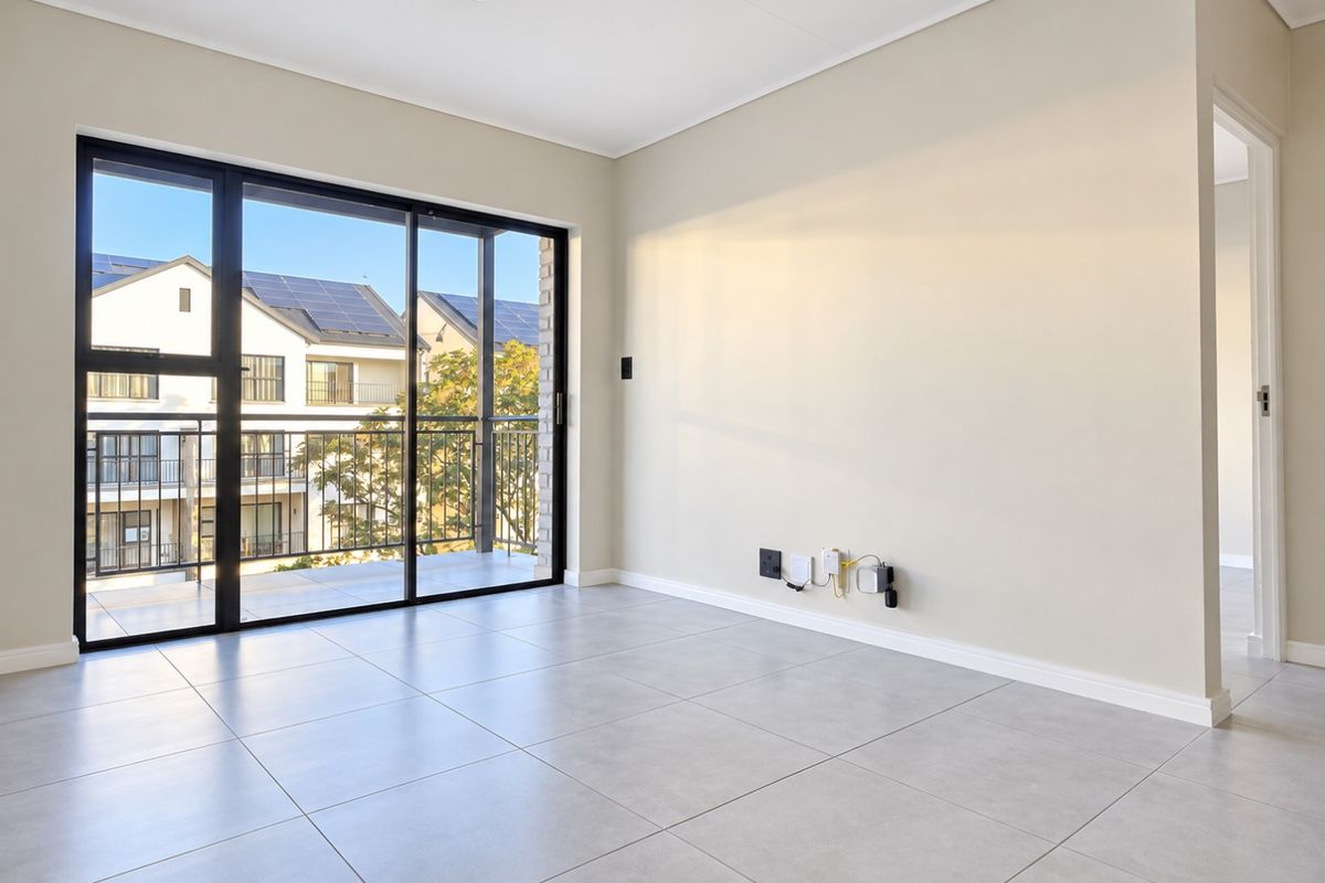 A bright, empty living room featuring large sliding glass doors leading to a balcony, neutral tiled flooring, and modern track lighting.