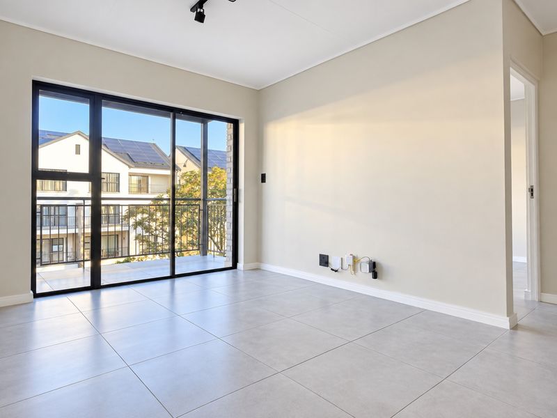 A bright, empty living room featuring large sliding glass doors leading to a balcony, neutral tiled flooring, and modern track lighting.