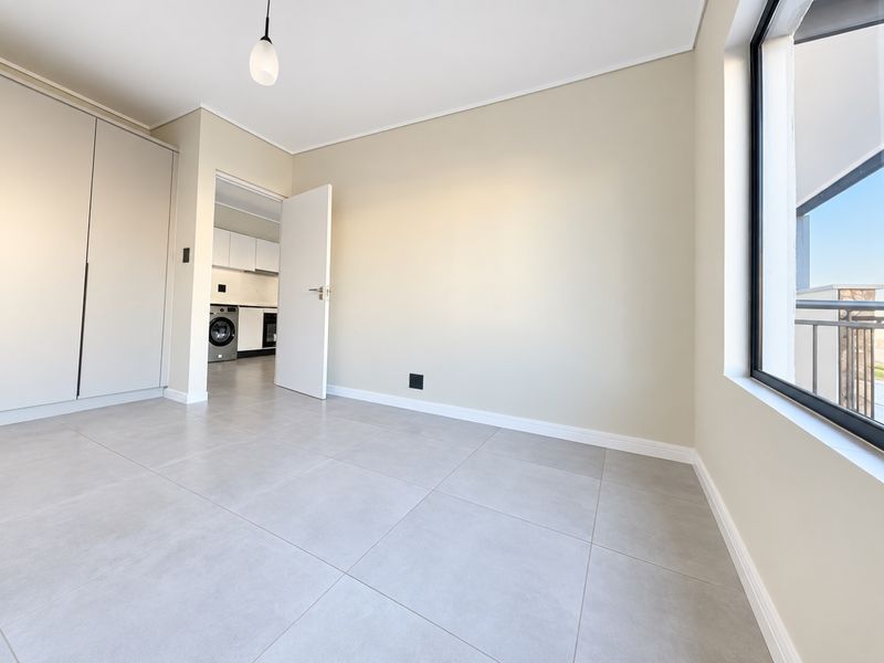 A bright, empty bedroom featuring neutral tiled flooring, built-in cupboards, and a large window with access to an adjacent kitchen area.