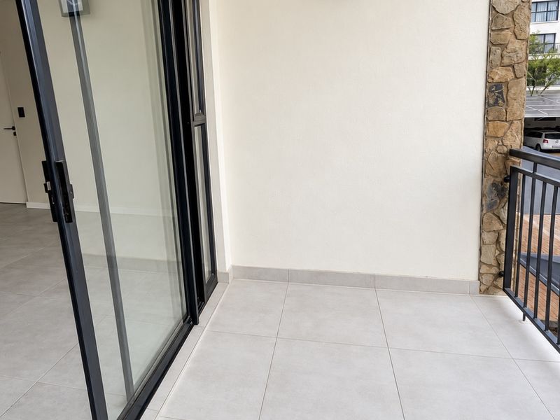 A clean, modern balcony featuring large format floor tiles, stone accent wall, and black framed sliding glass doors leading into the apartment.