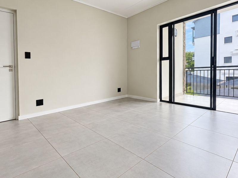 A bright, empty living area featuring modern neutral floor tiles, track lighting, and large sliding glass doors leading to a private balcony.