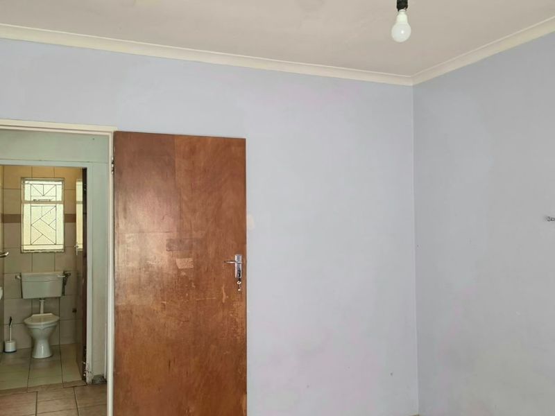 An empty bedroom featuring laminate flooring and a wooden door, with an adjacent bathroom visible. The room requires cosmetic updates to the walls and finishes.