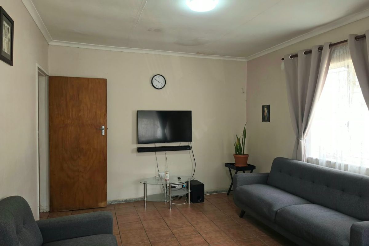 A living room featuring tiled flooring, coved ceilings, and a window with curtain fittings. The space shows signs of wear and ceiling water staining.