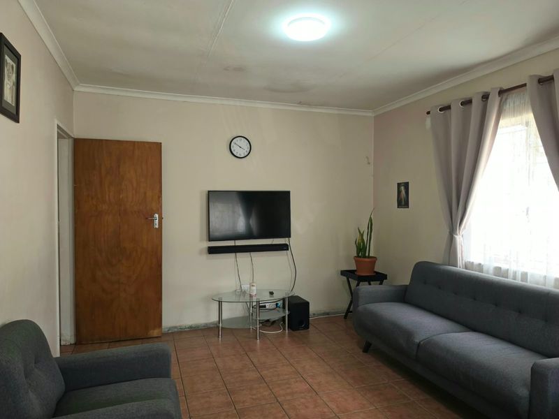 A living room featuring tiled flooring, coved ceilings, and a window with curtain fittings. The space shows signs of wear and ceiling water staining.