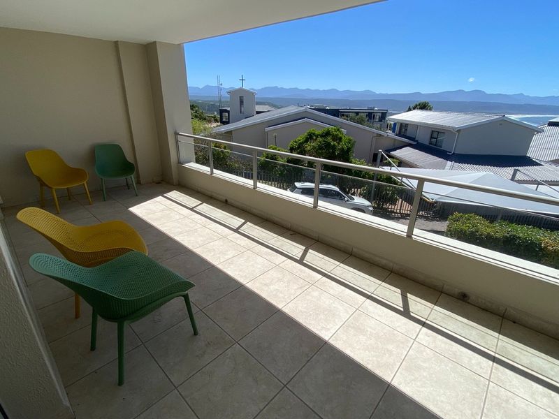 A spacious covered balcony featuring tiled flooring and a modern glass and metal balustrade, offering scenic views of the surrounding neighborhood and distant mountains.