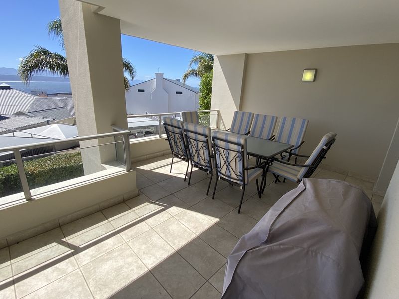 A spacious covered balcony featuring tiled flooring and a glass balustrade, offering an outdoor area suitable for dining with views of the surrounding neighborhood.