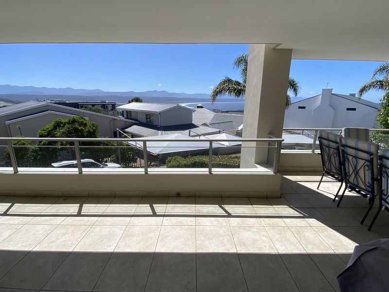 A spacious covered balcony featuring tiled flooring and a modern stainless steel and glass balustrade, offering expansive coastal and mountain views.