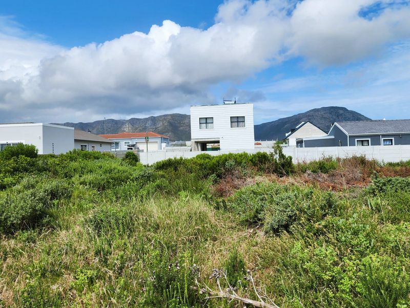 A vacant residential plot featuring overgrown vegetation and a precast concrete boundary wall, situated in a developed neighborhood with mountain views.