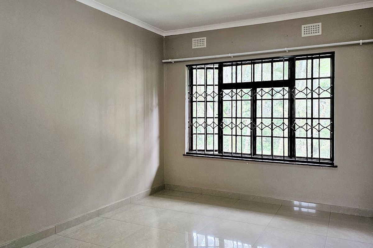 A bright, empty bedroom featuring neutral tiled flooring, large windows with security bars, and built-in ventilation grilles.