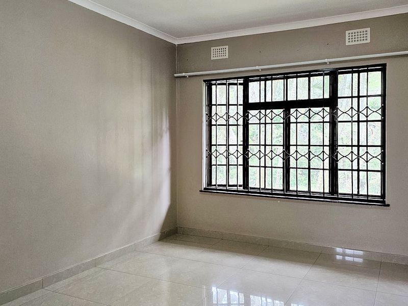 A bright, empty bedroom featuring neutral tiled flooring, large windows with security bars, and built-in ventilation grilles.
