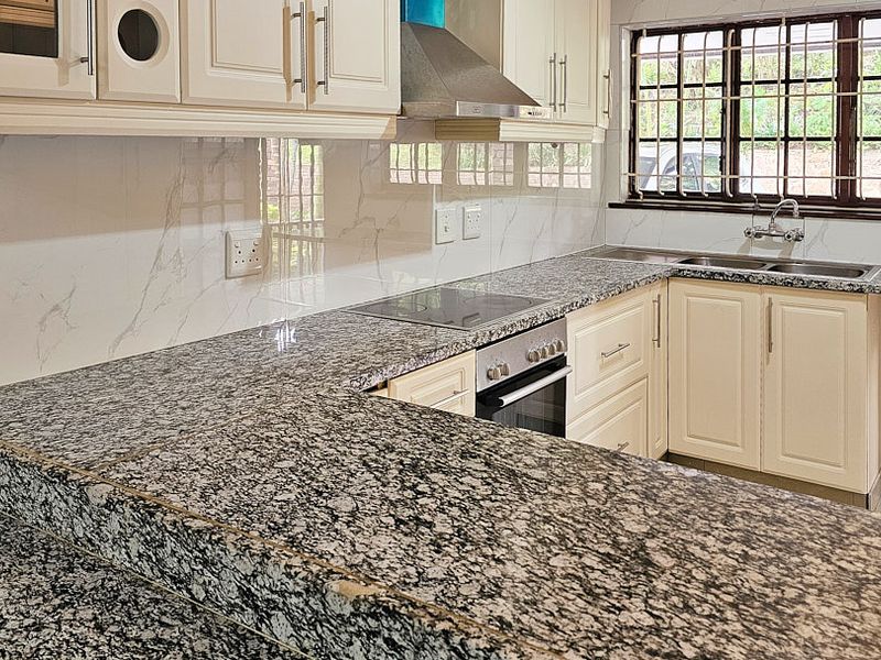 A functional kitchen featuring granite countertops, built-in oven, electric hob, and ample cabinetry, with a window providing natural light and security bars.