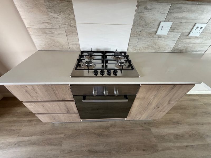 Modern kitchen section featuring a stainless steel gas hob, built-in oven, stone countertop, and wood-look laminate cabinetry with a tiled backsplash.
