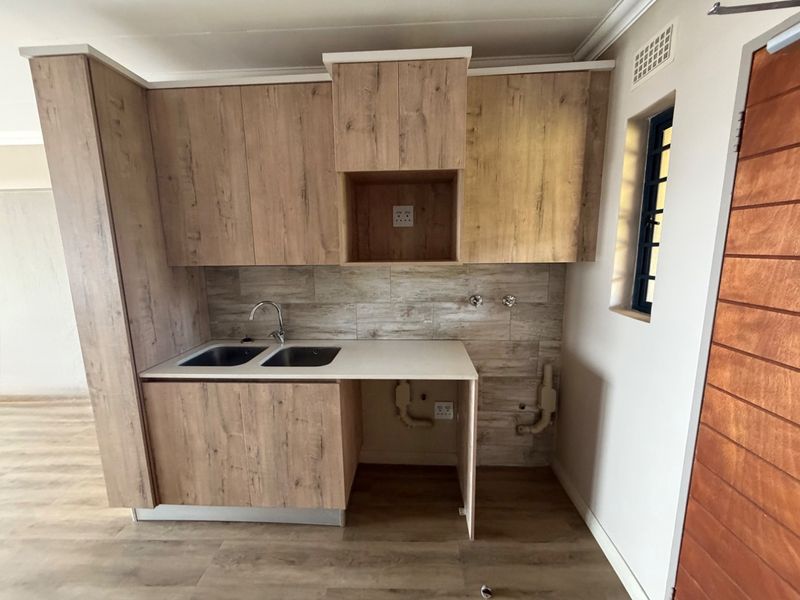 A compact kitchen featuring wood-look cabinetry, a double sink, tiled backsplash, and dedicated plumbing and electrical connections for appliances.