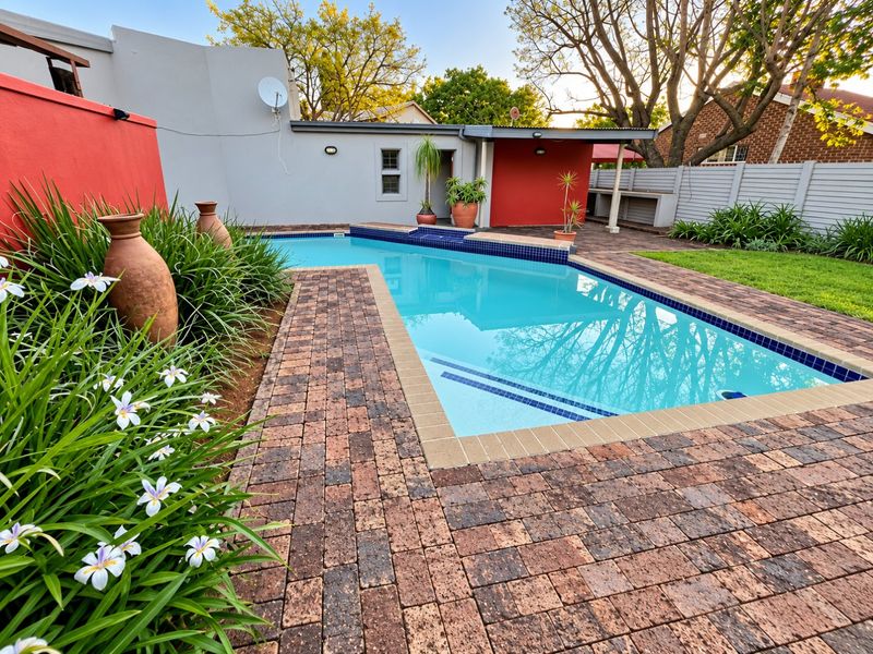 A well-maintained outdoor pool area featuring brick paving, a clear swimming pool, and a covered patio space in a landscaped garden setting.