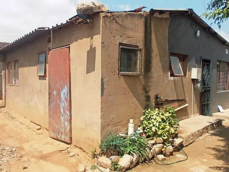 Rear view of a modest residential structure featuring plastered masonry walls, a corrugated metal roof, and basic security fixtures.