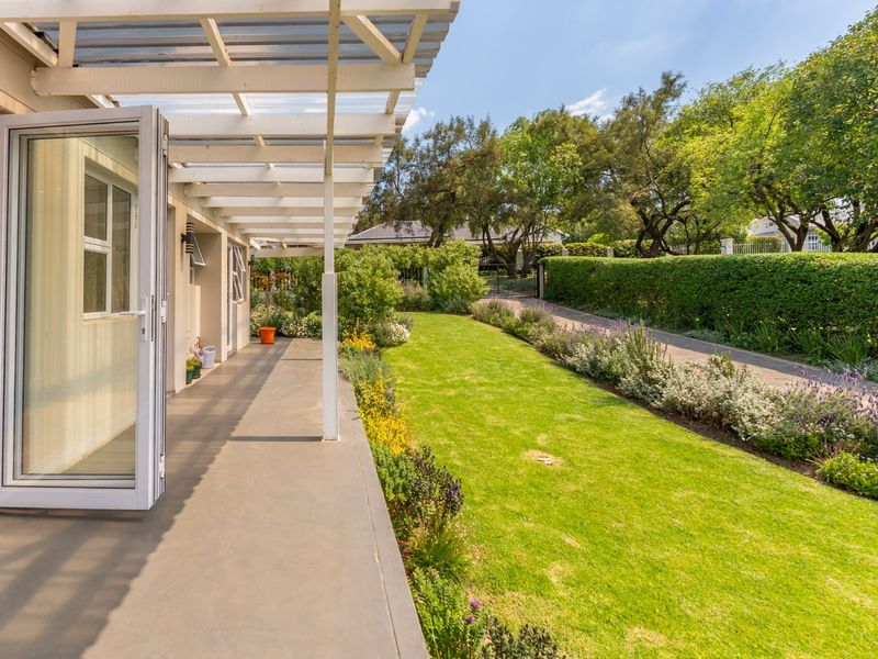 A well-maintained covered patio featuring a pergola, leading out to a lush, landscaped garden with a paved walkway and mature hedges.