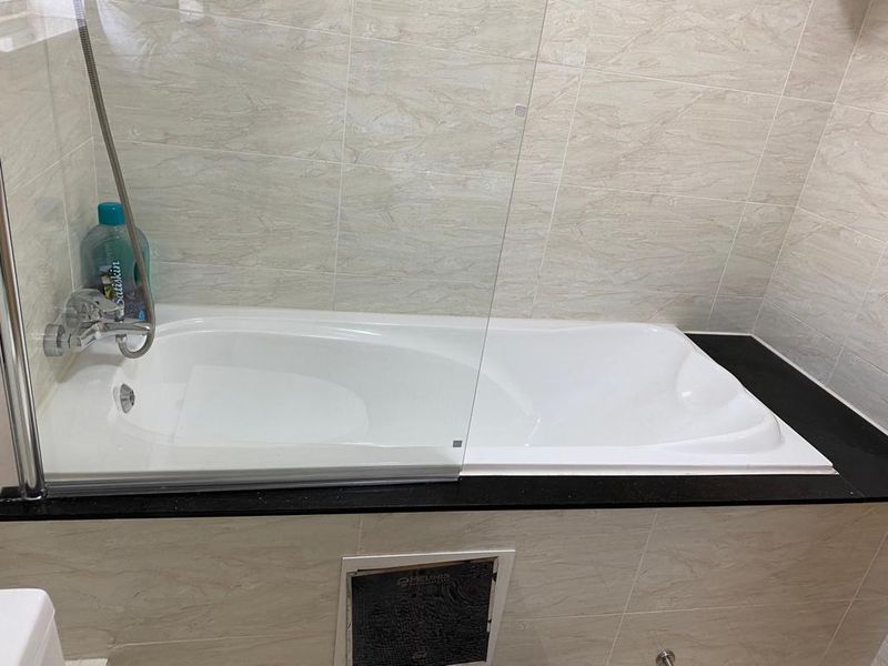 A clean, modern bathroom featuring a built-in bathtub with a glass shower screen, complemented by neutral marble-look wall tiles and a black granite surround.