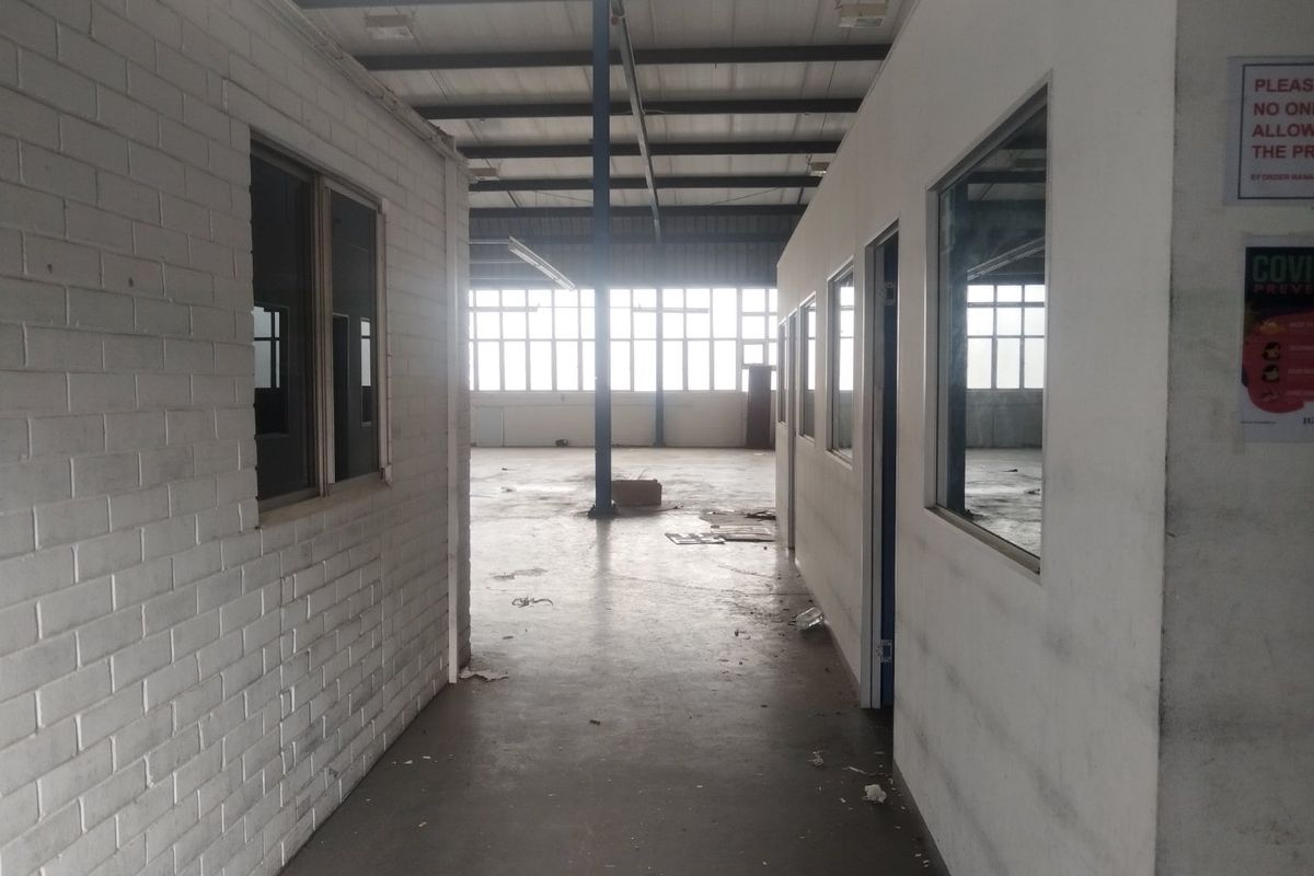 An industrial warehouse interior featuring partitioned office spaces, exposed steel roof trusses, and large windows providing natural light to the open floor area.