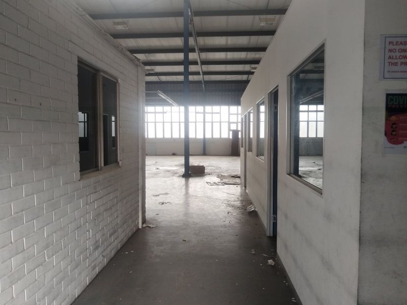 An industrial warehouse interior featuring partitioned office spaces, exposed steel roof trusses, and large windows providing natural light to the open floor area.