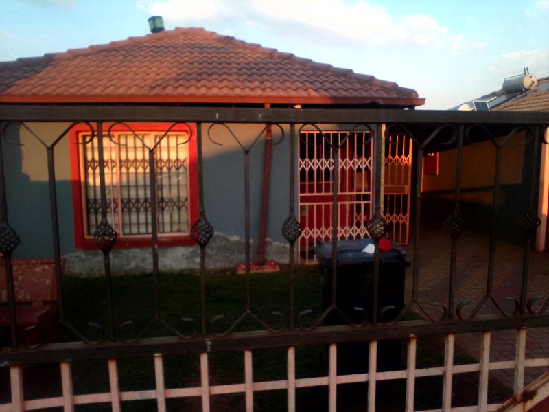 Front view of a single-story house featuring a tiled roof, security gates, and a paved driveway, showing signs of exterior wall wear.