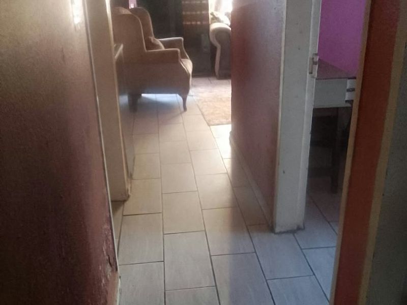 A narrow hallway featuring tiled flooring, leading into an adjacent living area with visible wall wear.