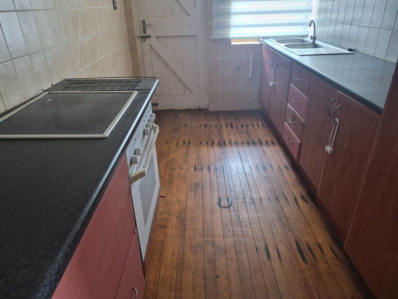 A functional kitchen featuring built-in cabinetry, laminate countertops, an electric stove, and a stainless steel sink, with original timber flooring throughout.