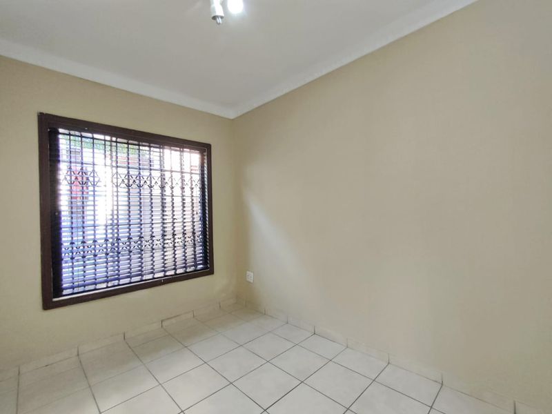 A clean, empty bedroom featuring neutral tiled flooring, coving, and a window fitted with security burglar bars.
