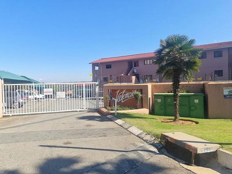 Front entrance view of the Valencia townhouse complex featuring a secure gated access point, perimeter wall with electric fencing, and paved driveway.