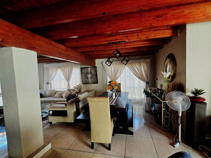 Spacious open-plan living and dining area featuring warm exposed timber ceiling beams, large windows with natural light, and neutral tiled flooring throughout.