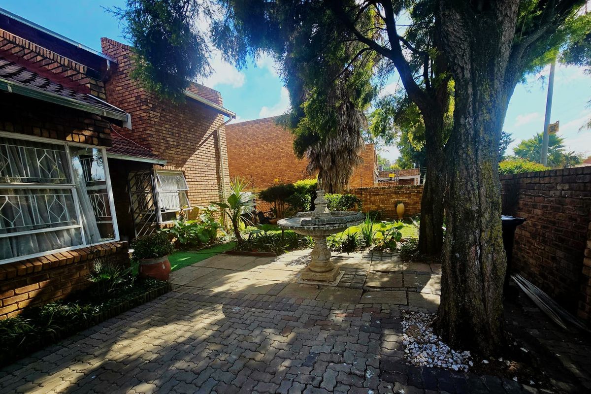 A private rear garden featuring a brick-faced building, paved patio area, and a central stone fountain surrounded by mature trees and boundary walls.