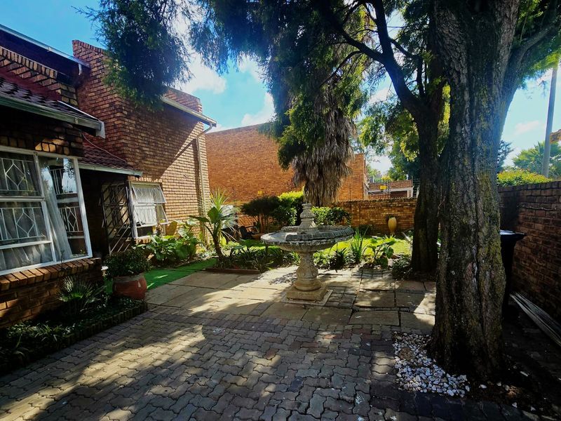 A private rear garden featuring a brick-faced building, paved patio area, and a central stone fountain surrounded by mature trees and boundary walls.