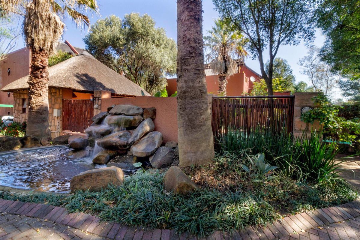 A landscaped rear garden area featuring a stone water feature, mature trees, and brick paving, adjacent to a building with a thatched roof.