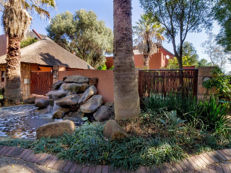 A landscaped rear garden area featuring a stone water feature, mature trees, and brick paving, adjacent to a building with a thatched roof.
