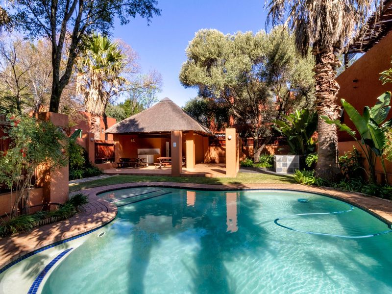 A private outdoor pool area featuring a thatched lapa with a built-in braai, surrounded by established trees and landscaped garden space.