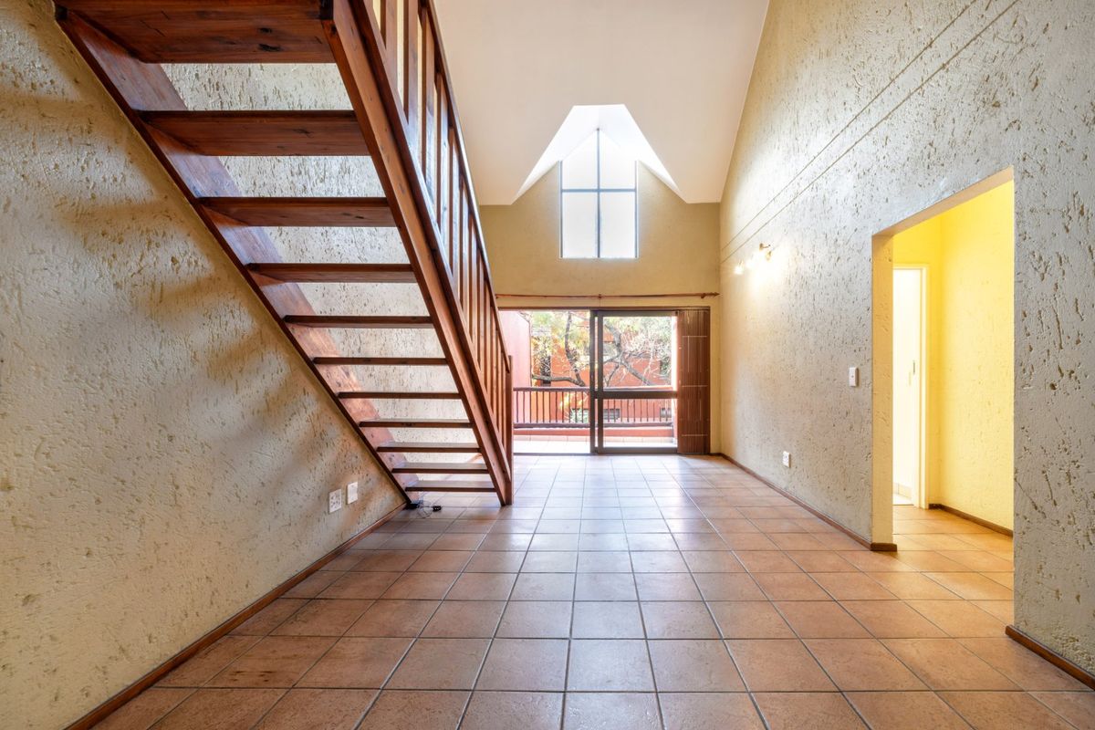 An empty, open-plan living area featuring a wooden staircase, high vaulted ceilings, tiled floors, and a sliding door leading to a balcony.