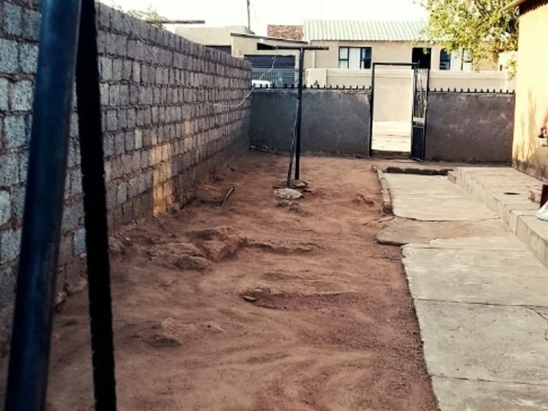 A rear yard area featuring a concrete boundary wall, a metal gate, and a concrete pathway, currently with an unlandscaped dirt surface.