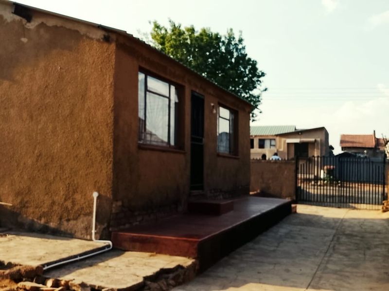 Front view of a single-story house featuring a concrete driveway, perimeter wall with security gate, and a raised concrete porch entrance.