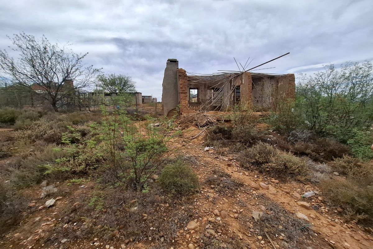 A view of a dilapidated, partially collapsed brick structure situated on a vacant residential plot with overgrown vegetation.