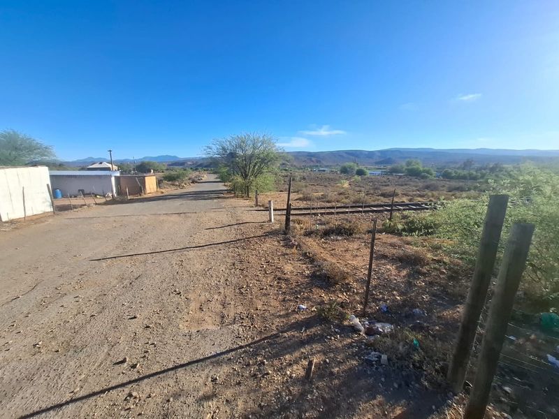 A view of an unpaved road and surrounding vacant land with wire fencing and distant mountain views.