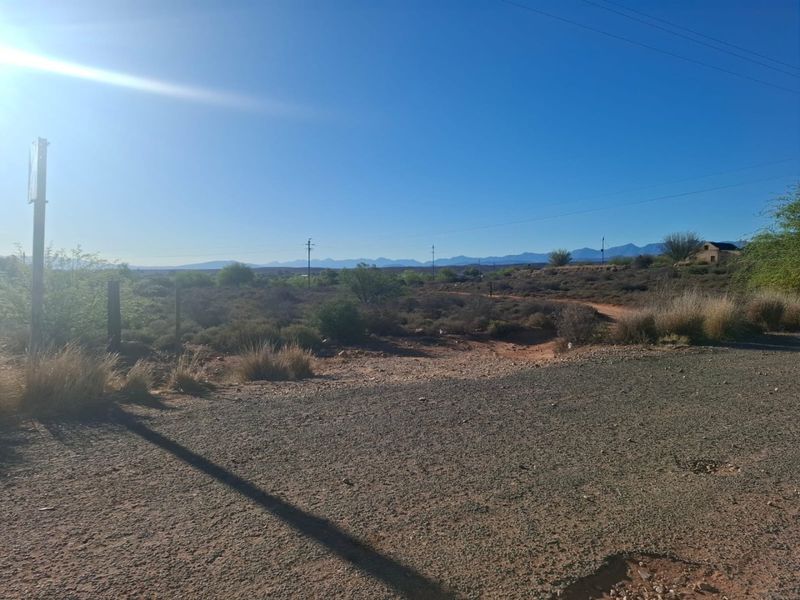 A wide-angle view of the vacant residential land featuring natural landscape, gravel access, and distant mountain views under a clear sky.