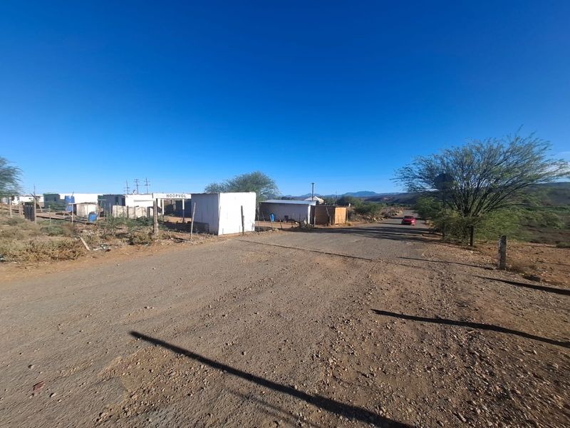 A street-level view of a rural residential area featuring unpaved roads, basic structures, and surrounding natural landscape.