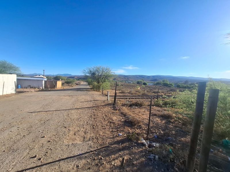 A view of an unpaved road and surrounding vacant land with wire fencing and mountain views in the distance.
