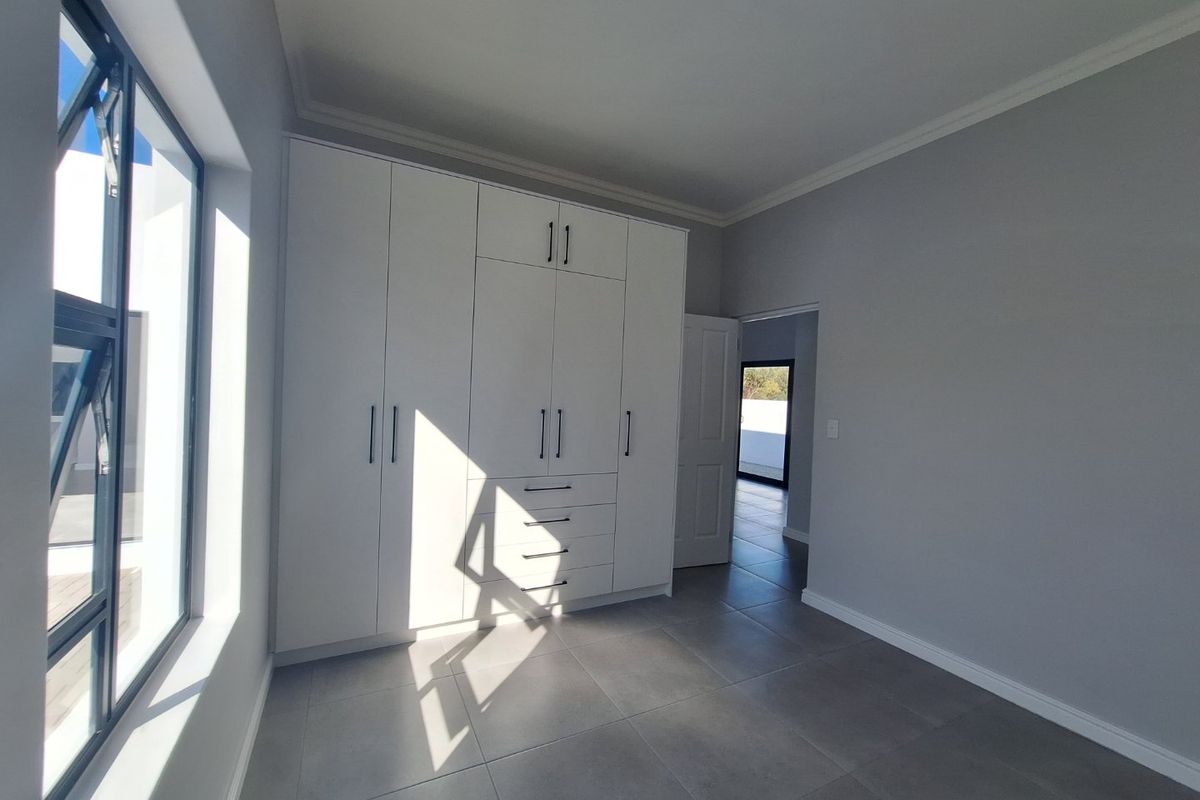 A bright, modern bedroom featuring neutral grey tiled flooring, crisp white walls, and extensive built-in cupboard storage with contemporary hardware.