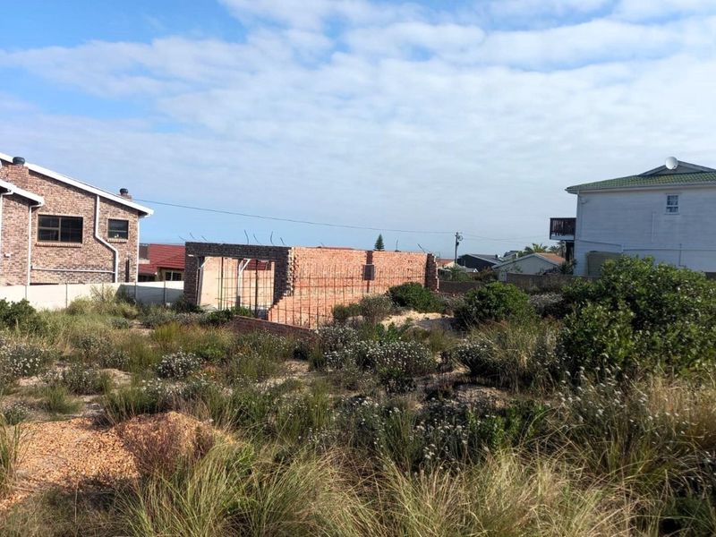 A vacant residential plot featuring partially constructed brick walls and exposed steel reinforcement, surrounded by natural coastal vegetation.
