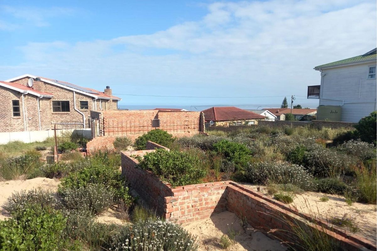 A vacant residential plot featuring unfinished brick wall structures and overgrown natural vegetation, situated in a developed coastal neighborhood.