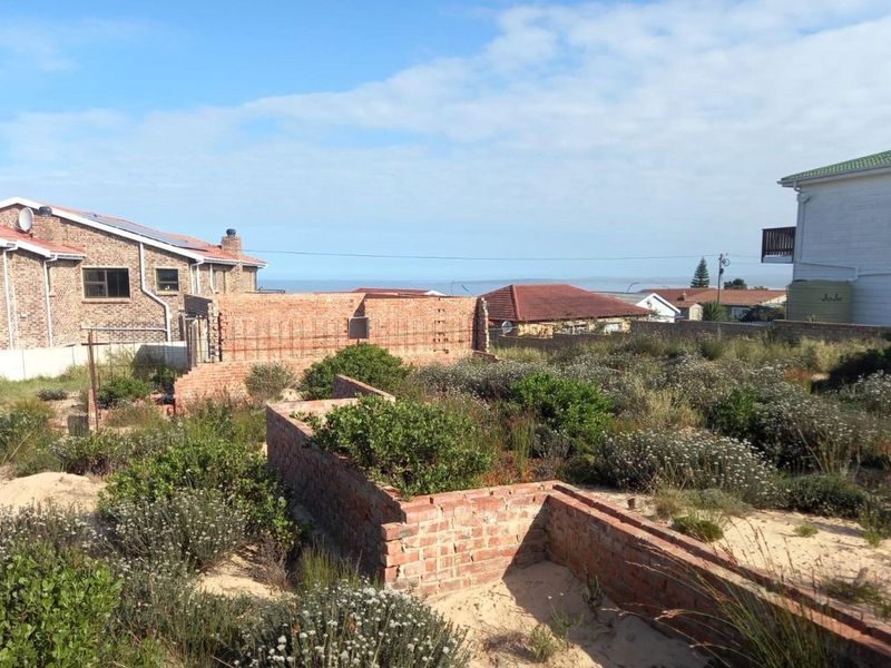A vacant residential plot featuring unfinished brick wall structures and overgrown natural vegetation, situated in a developed coastal neighborhood.